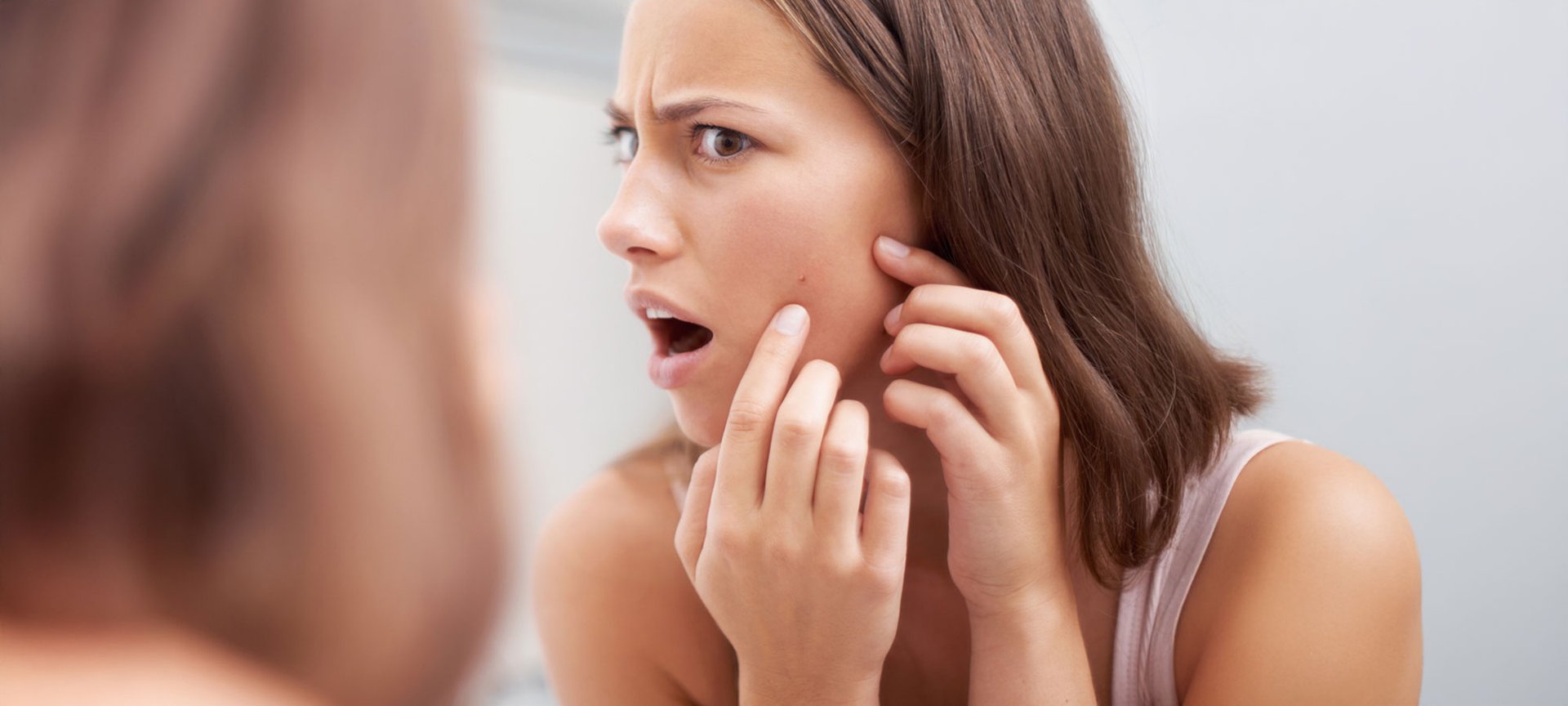 Frau sieht im Spiegel einen Pickel in ihrem Gesicht, zeigt darauf und schaut unglücklich.