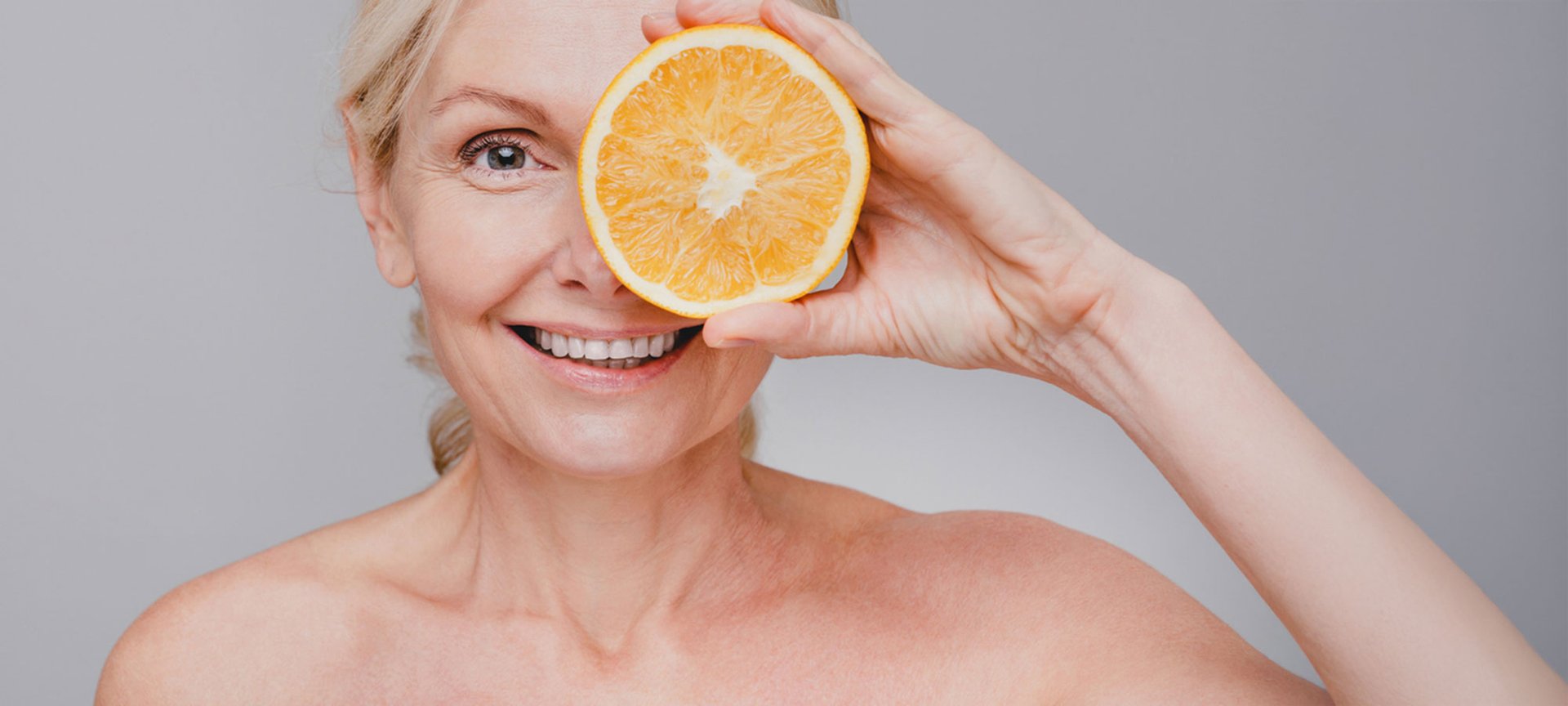 Eine ältere Frau mit blonden Haaren guckt lächelnd in die Kamera und hält sich mit der linken Hand eine Orangenscheibe vor das linke Auge.