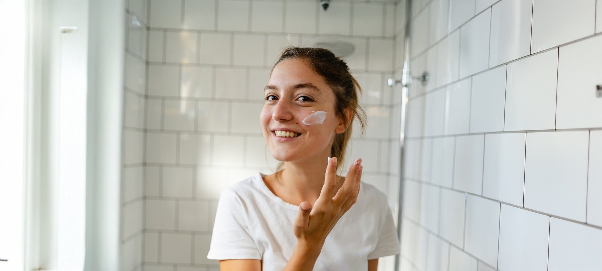 Eine junge Frau steht lächelnd im Badezimmer und trägt mit ihrer rechten Hand Creme auf ihre linke Wange auf.