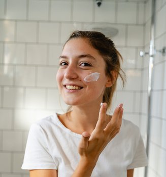 Eine junge Frau steht lächelnd im Badezimmer und trägt mit ihrer rechten Hand Creme auf ihre linke Wange auf.