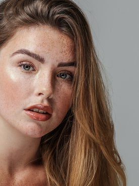 Junge Frau mit Sommersprossen und langen, glatten, dunkelblonden Haaren sieht in die Kamera