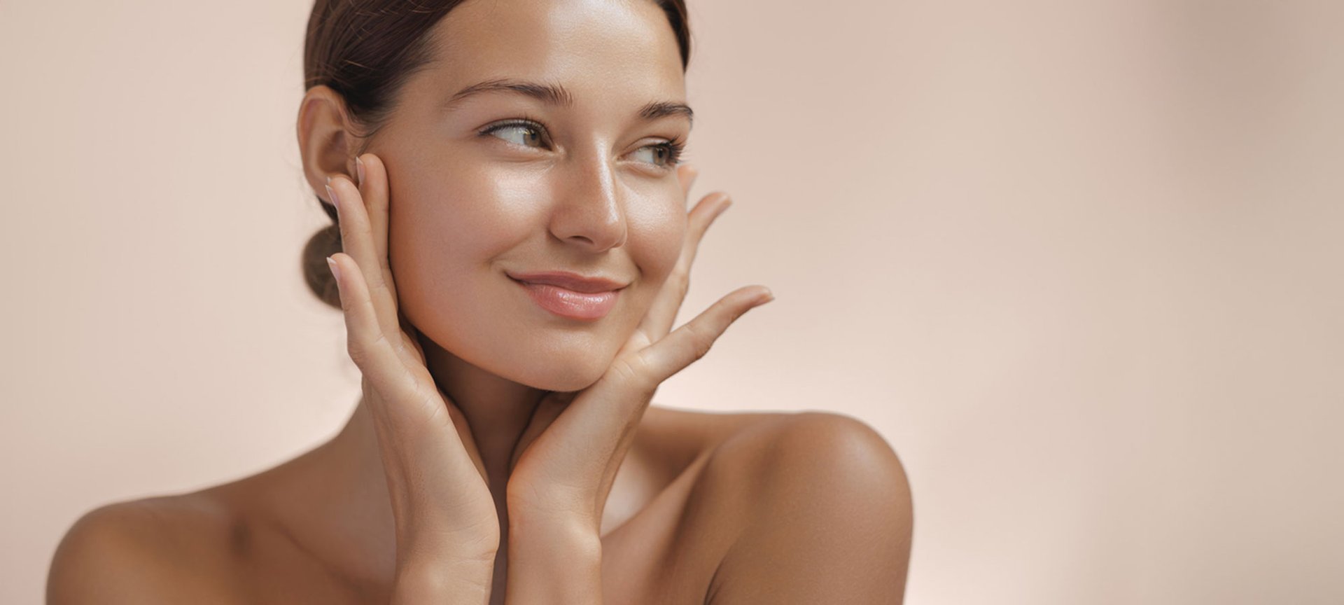 Gesicht und Schultern einer jungen Brünetten von vorne. Sie blickt lächelnd nach links und hält beide Hände an ihre Wangen.