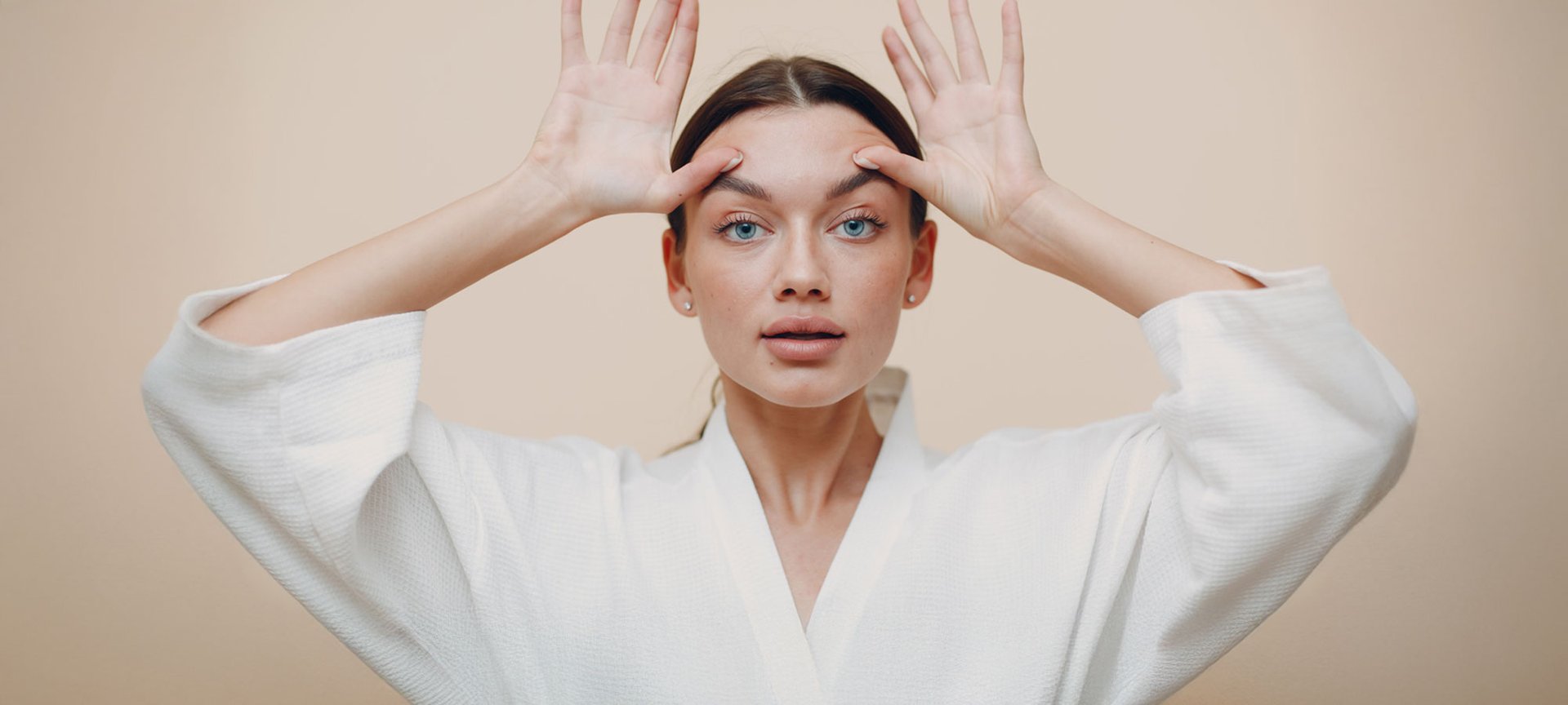 Eine Brünette mit blauen Augen blickt in die Kamera. Sie hält beide Hände in Stirnhöhe, mit den Handflächen nach vorne, dabei drückt sie mit den Daumen ihre Augenbrauen nach oben