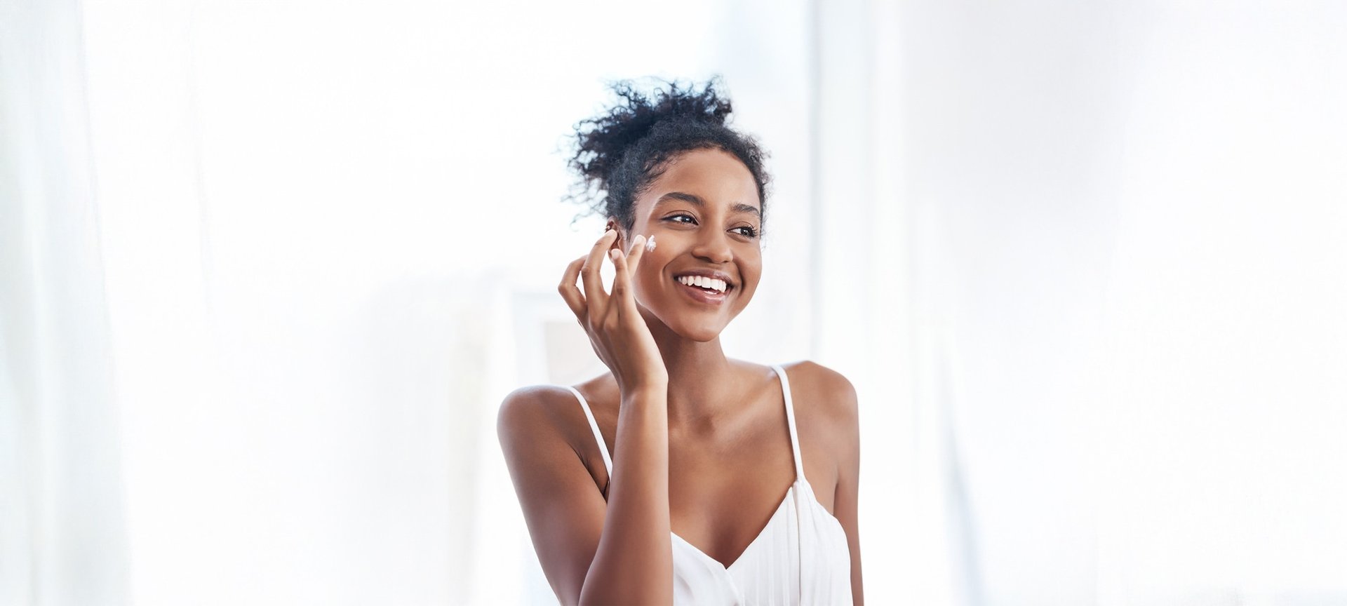 Eine Frau mit hochgesteckten Haaren tupft sich lächelnd Creme auf die rechte Wange.