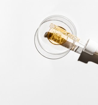 Eine Petrischale über der eine Pipette liegt, vor einem weißen Hintergrund. In der Pipette und im Zentrum der Schale ist ein goldgelbes Serum mit viele kleinen Bläschen.