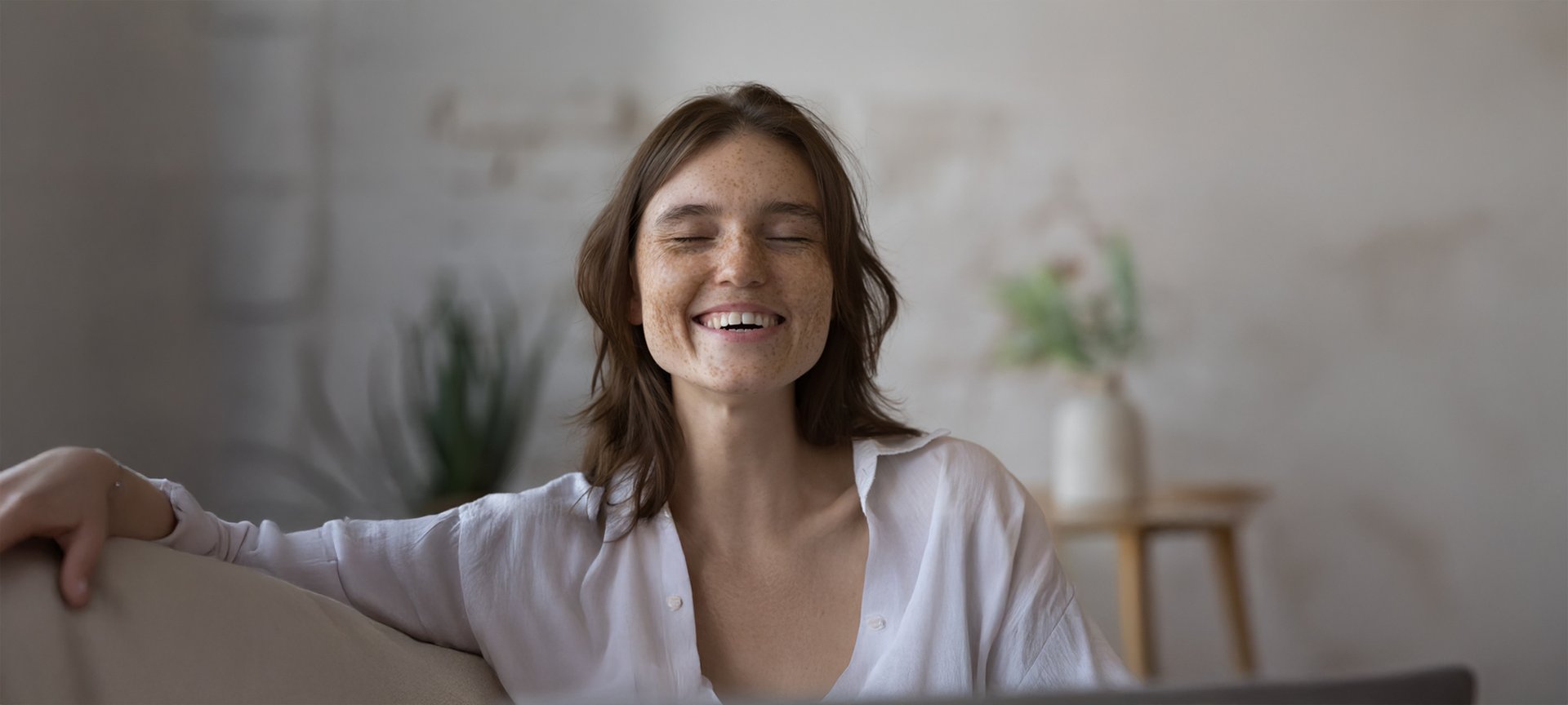 Frontalansicht einer jungen Frau mit vielen Sonnenflecken im ganzen Gesicht und braunen Haaren. Sie hat die Augen geschlossen und lächelt.