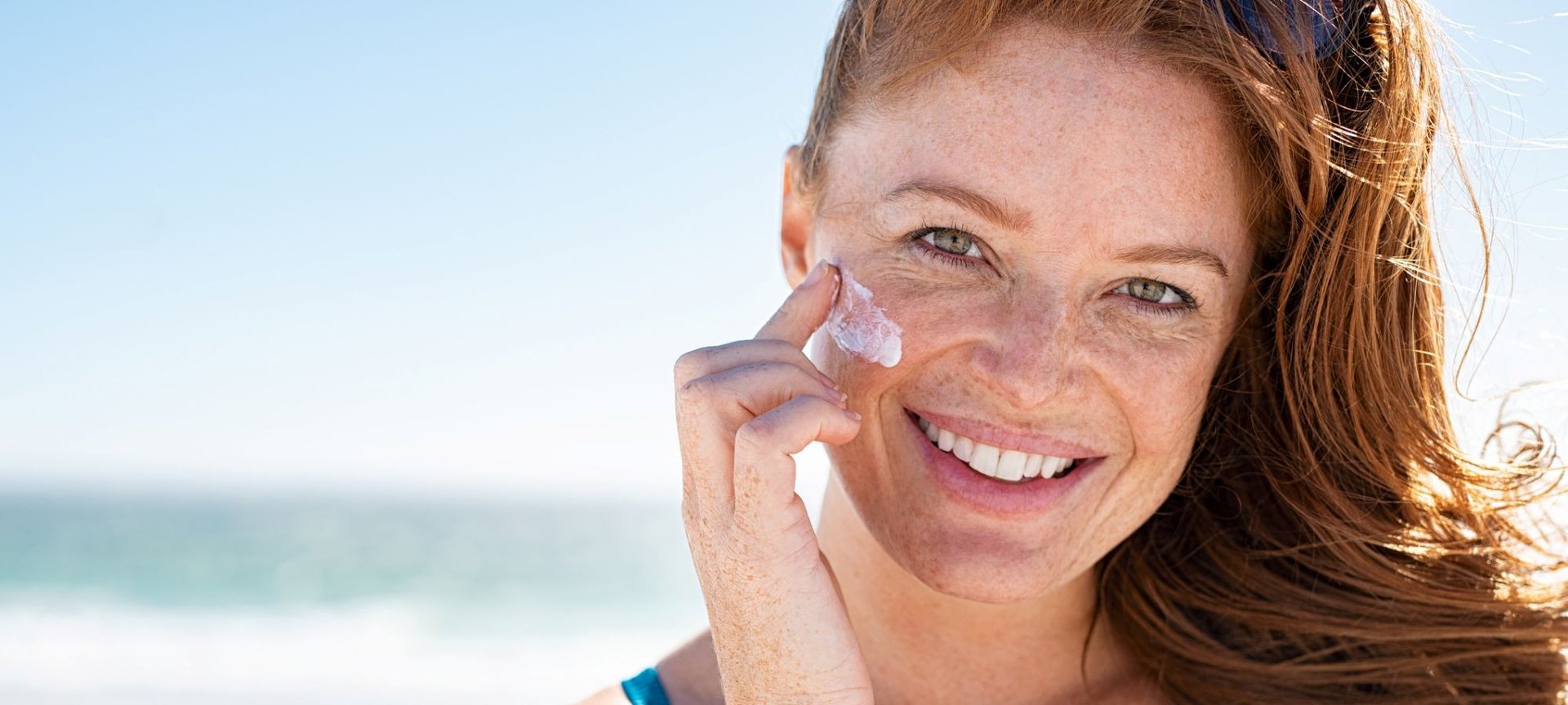 Eine lächelnde Frau mittleren Alters trägt am Strand Sonnencreme auf ihr Gesicht auf