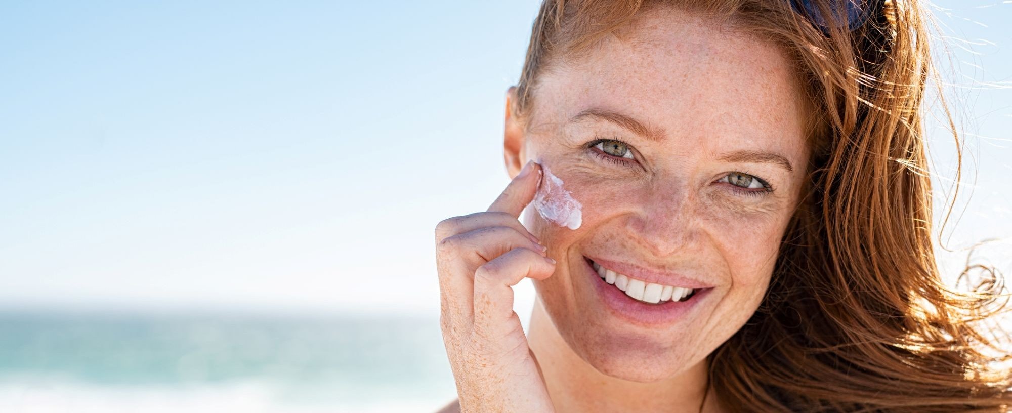 Eine lächelnde Frau mittleren Alters trägt am Strand Sonnencreme auf ihr Gesicht auf