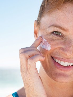 Eine lächelnde Frau mittleren Alters trägt am Strand Sonnencreme auf ihr Gesicht auf
