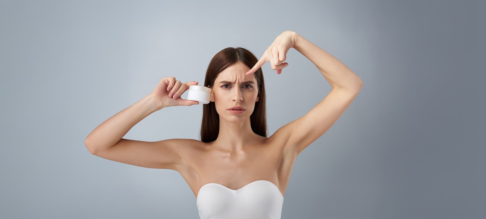 Eine junge braunhaarige Frau guckt erbost in die Kamera. Dabei hält sie mit der rechten Hand einen Cremetiegel auf Gesichtshöhe und zeigt mit dem linke Zeigefinger auf die Zornesfalten auf ihrer Stirn.