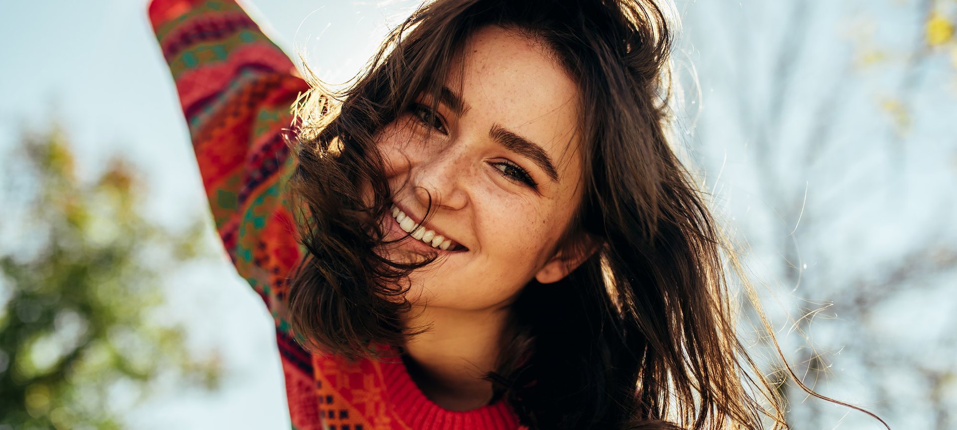 Eine Frau lächelt in die Kamera, sie trägt einen herbstlichen Pullover hinter ihr sind Bäume