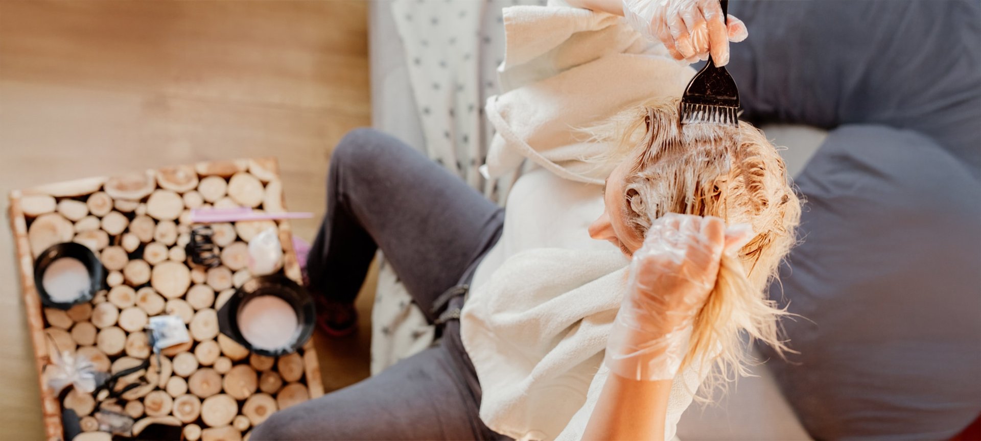 Eine blonde Frau von schräg oben. Sie sitzt auf einem Sofa. Vor sich auf dem Tisch steht eine Schüssel mit Blondierung. Sie hat ein Handtuch über den Schultern, durchsichtige Handschuhe an und trägt mit einem schwarzen Pinsel die Blondierung auf ihre Haare auf.