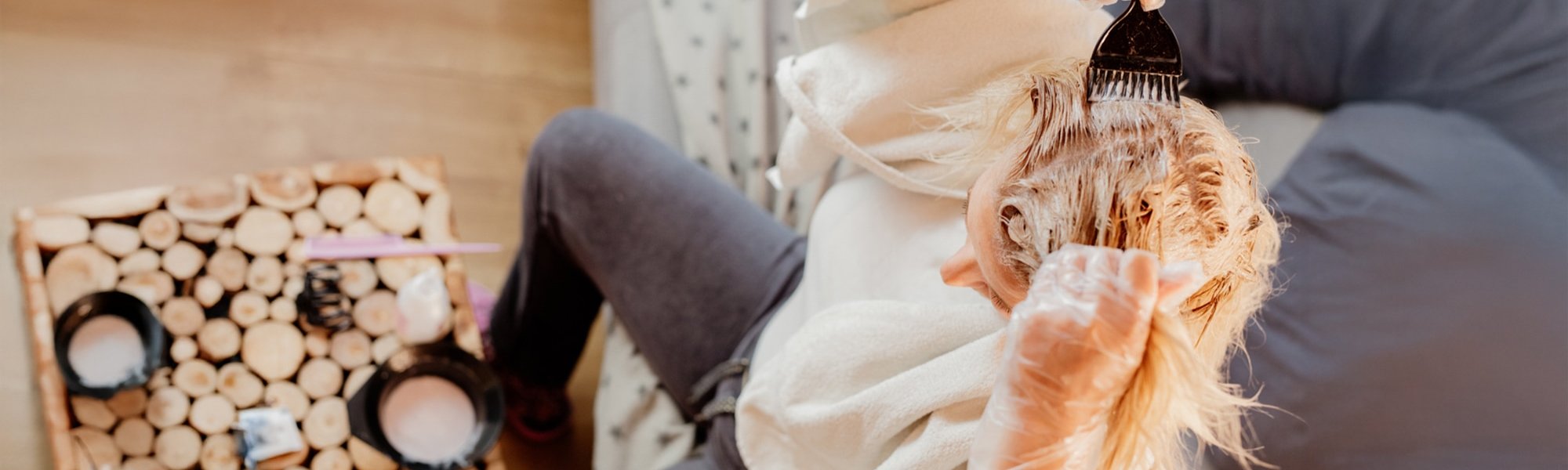 Eine blonde Frau von schräg oben. Sie sitzt auf einem Sofa. Vor sich auf dem Tisch steht eine Schüssel mit Blondierung. Sie hat ein Handtuch über den Schultern, durchsichtige Handschuhe an und trägt mit einem schwarzen Pinsel die Blondierung auf ihre Haare auf.