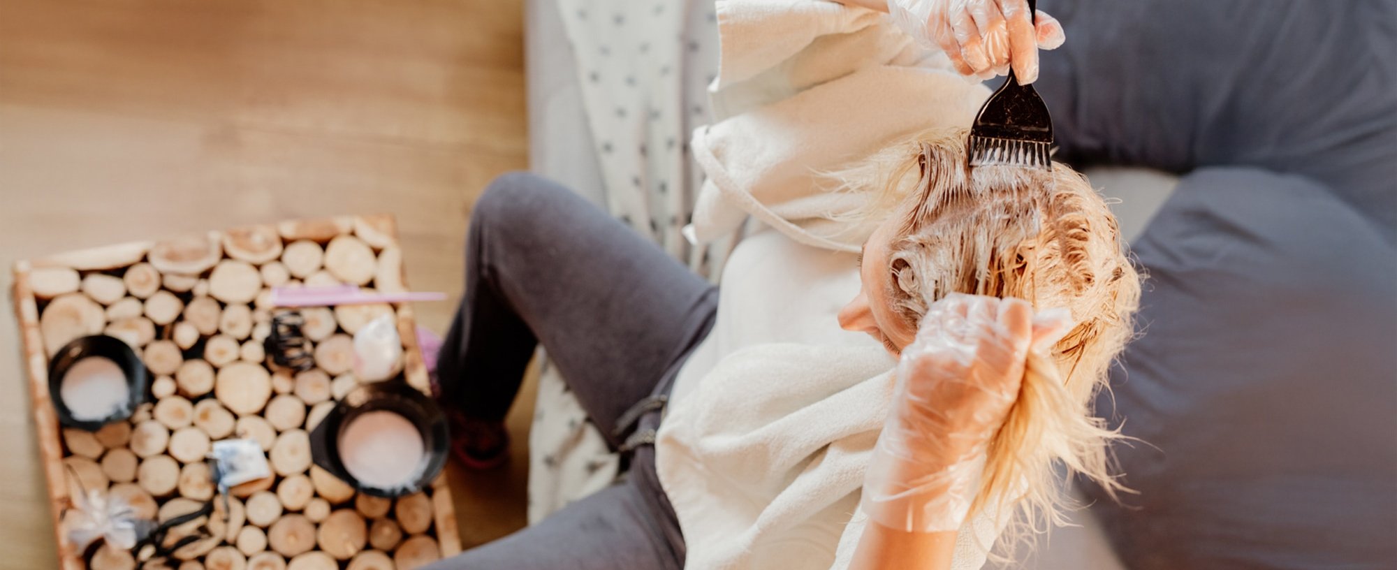 Eine blonde Frau von schräg oben. Sie sitzt auf einem Sofa. Vor sich auf dem Tisch steht eine Schüssel mit Blondierung. Sie hat ein Handtuch über den Schultern, durchsichtige Handschuhe an und trägt mit einem schwarzen Pinsel die Blondierung auf ihre Haare auf.