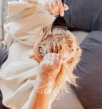 Eine blonde Frau von schräg oben. Sie sitzt auf einem Sofa. Vor sich auf dem Tisch steht eine Schüssel mit Blondierung. Sie hat ein Handtuch über den Schultern, durchsichtige Handschuhe an und trägt mit einem schwarzen Pinsel die Blondierung auf ihre Haare auf.