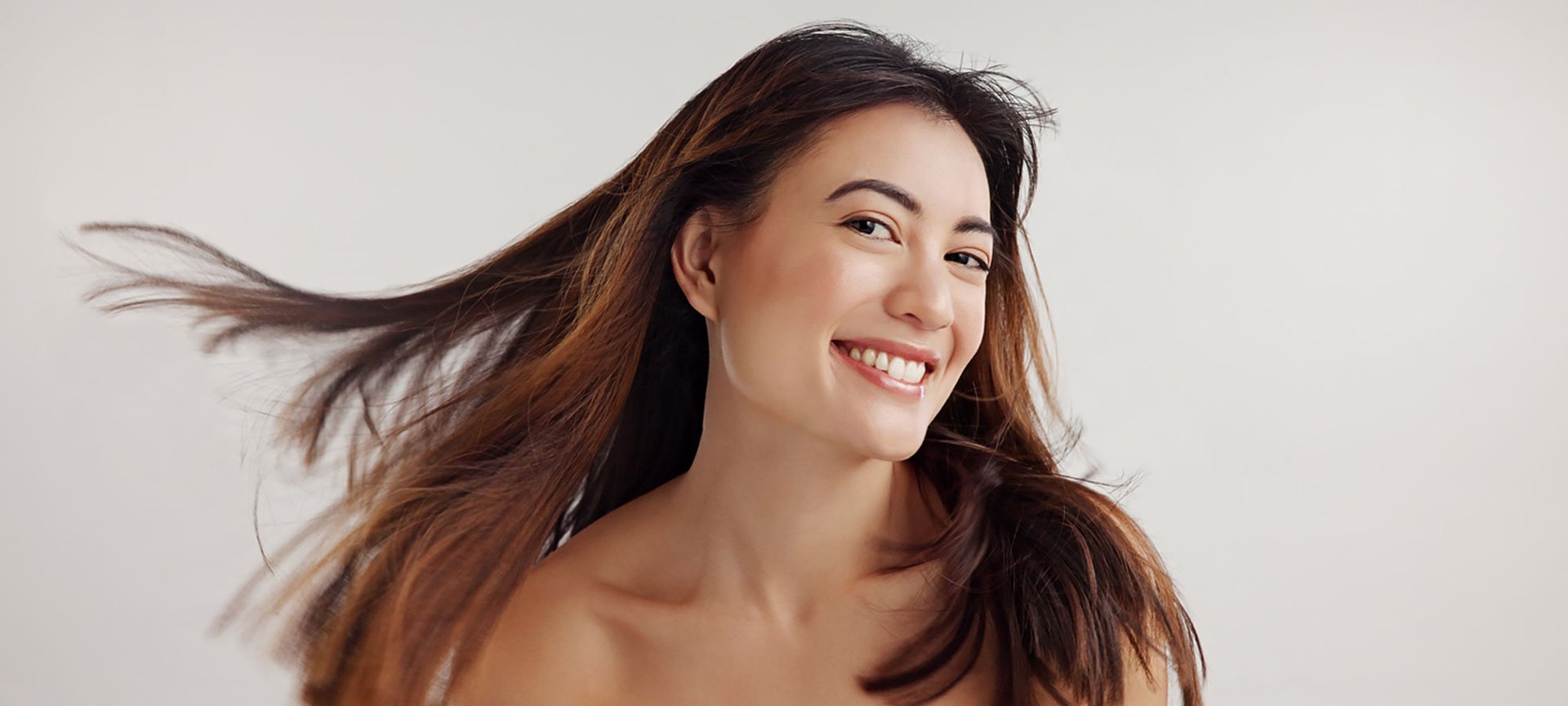 Junge Frau mit braunem Haar lächelt vor grauem Hintergrund.