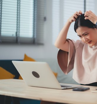 Junge Frau vor einem Laptop kratzt sich wegen trockener Kopfhaut am Kopf