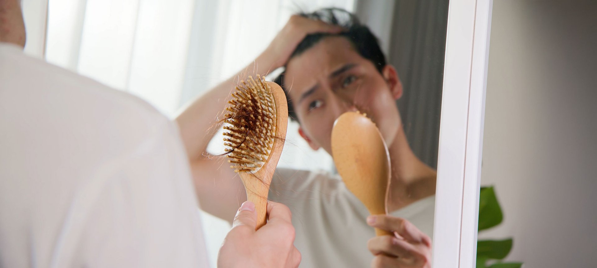 Besorgter junger Mann mit Haarausfall schaut in den Spiegel und auf seine Haarbürste