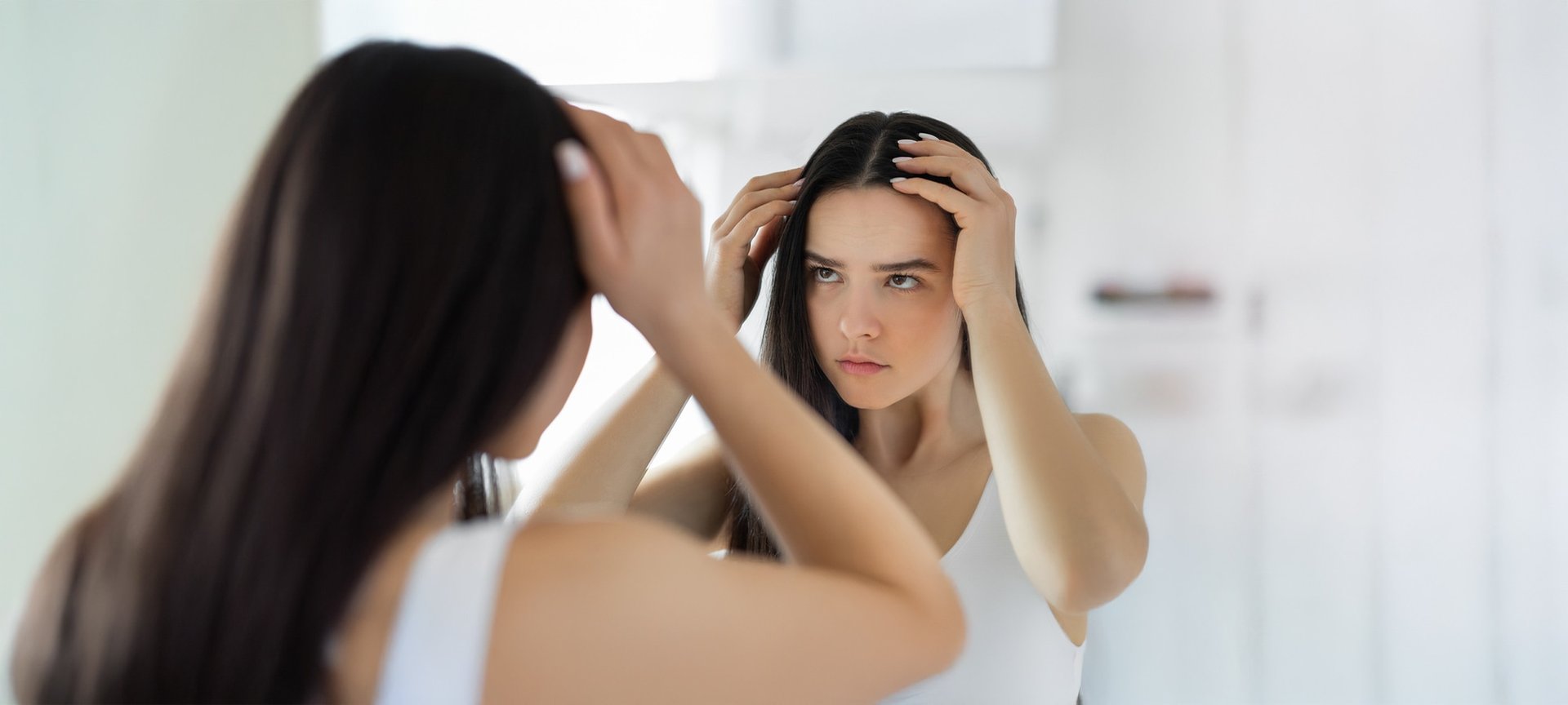 Junge Frau sieht sich im Spiegel unglücklich ihre Kopfhaut an.