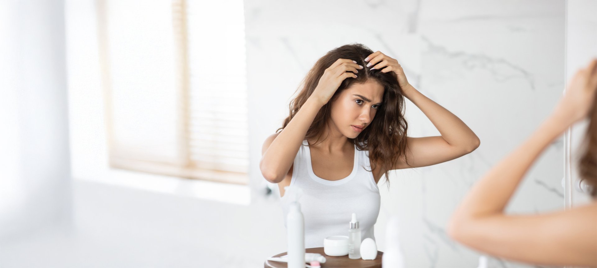 Eine junge Frau mit langen braunen Haaren befühlt ihren Haaransatz und blickt dabei kritisch in einen Wandspiegel.