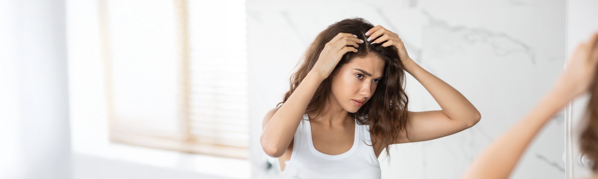 Eine junge Frau mit langen braunen Haaren befühlt ihren Haaransatz und blickt dabei kritisch in einen Wandspiegel.