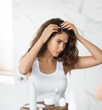 Eine junge Frau mit langen braunen Haaren befühlt ihren Haaransatz und blickt dabei kritisch in einen Wandspiegel.