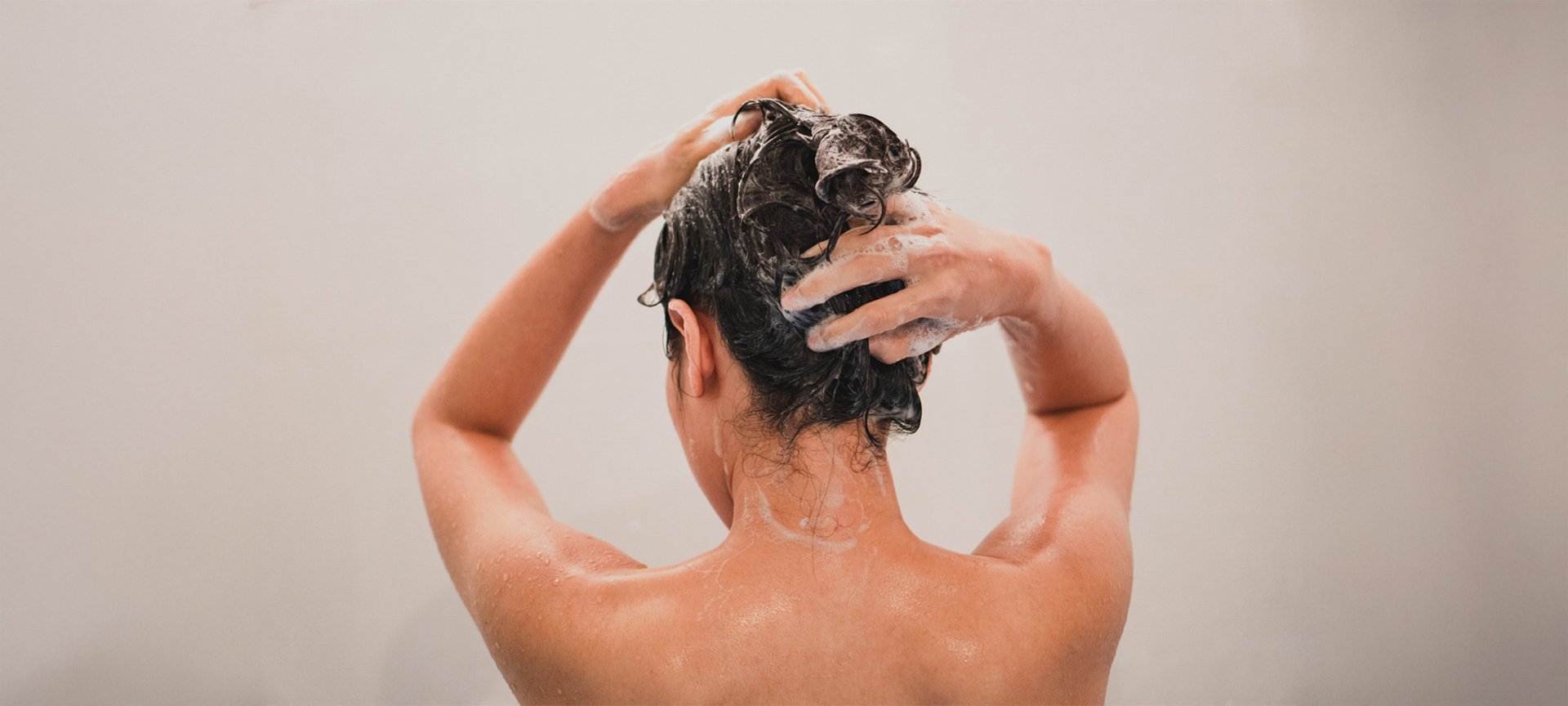 Eine brünette Frau von hinten, die sich mit beiden Händen die Haare wäscht.
