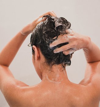 Eine brünette Frau von hinten, die sich mit beiden Händen die Haare wäscht.