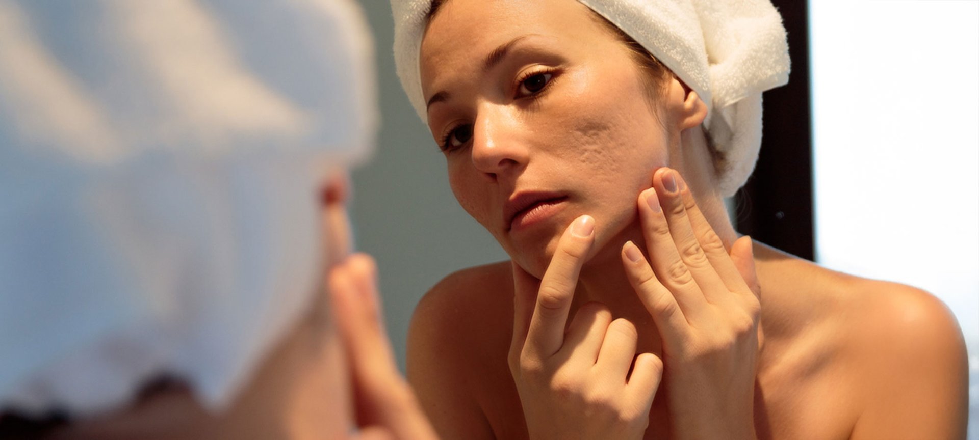 Blick über die Schulter einer Frau mit Handtuchturban, die im Spiegel die Narben auf ihrer linken Wange betrachtet und mit den Hände ihr Gesicht befühlt.