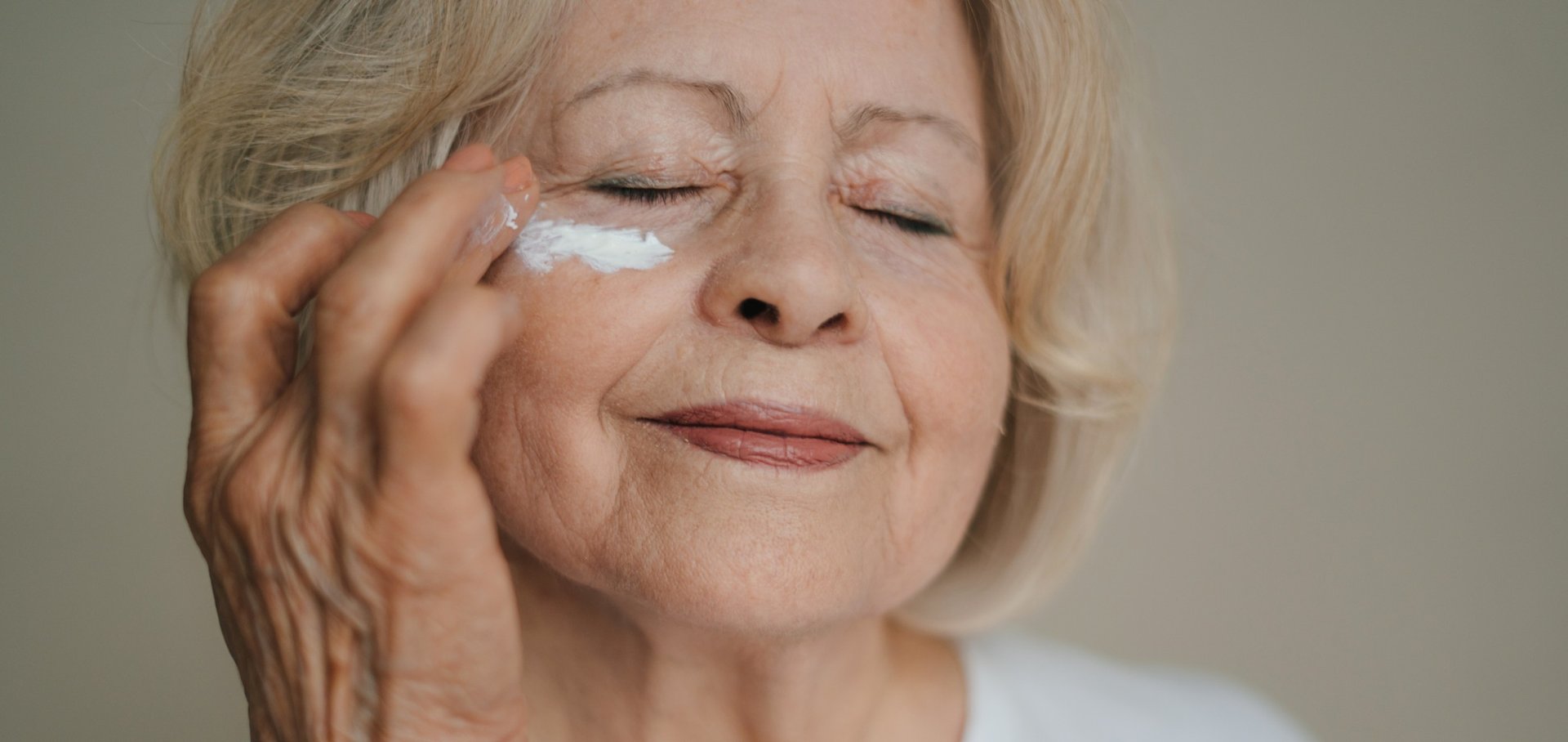 Eine ältere Dame mit geschlossenen Augen trägt Augencreme unter ihrem Auge auf