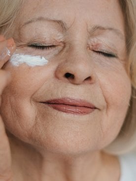 Eine ältere Dame mit geschlossenen Augen trägt Augencreme unter ihrem Auge auf
