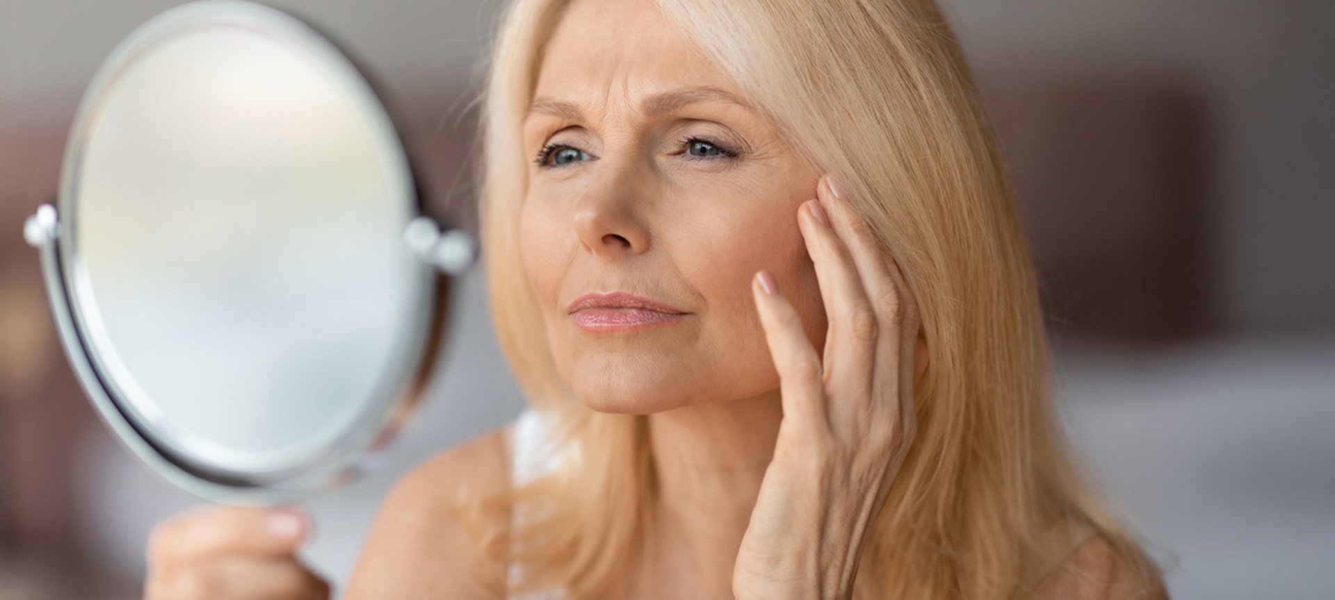 Frau mit blonden Haaren betrachtet kritisch ihre Gesichtsfalten in einem runden Kosmetikspiegel