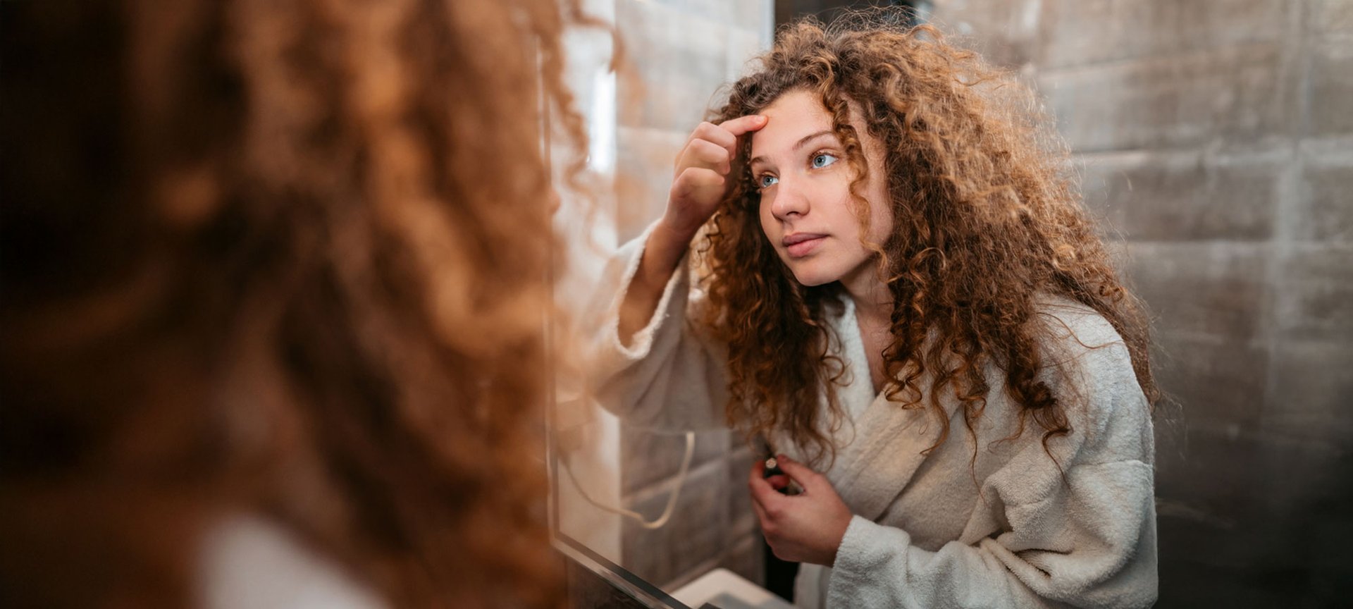 Junge Frau mit braunen Locken beäugt im Spiegel einen Pickel auf ihrer Stirn