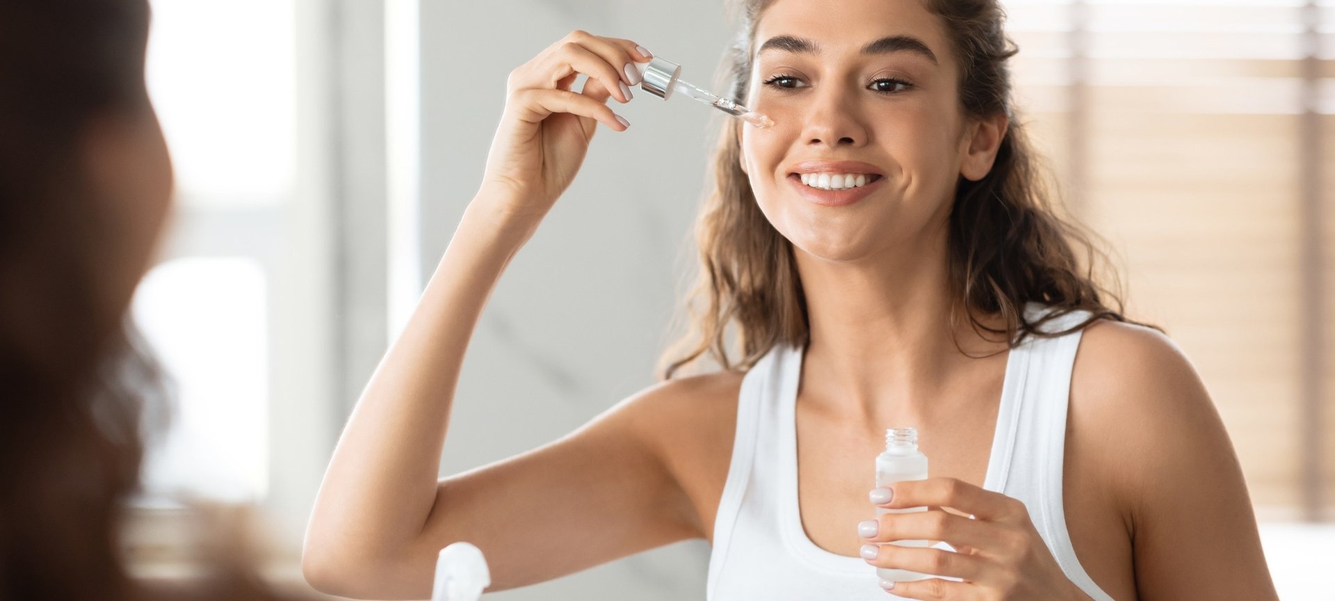 Junge Frau steht vor einem Spiegel trägt ein Feuchtigkeitserum auf ihre Wangen auf