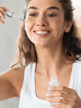 Junge Frau steht vor einem Spiegel trägt ein Feuchtigkeitserum auf ihre Wangen auf