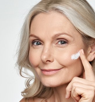 Eine lächelnde, ältere Dame mit Falten trägt Sonnencreme auf ihre Wange auf