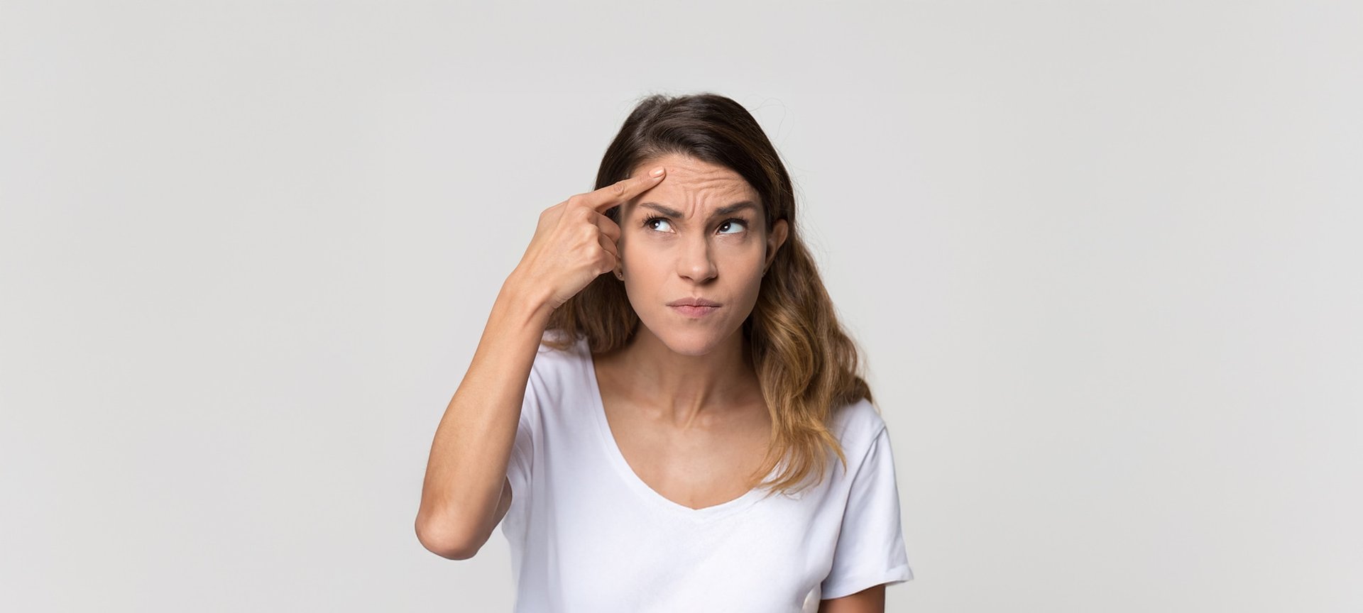 Eine braunhaarige Frau befühlt mit ihrem rechten Zeigefinger ihre Stirn, auf der viele Falten zu sehen sind. Dabei guckt sie skeptisch nach oben, in Richtung ihres Zeigefingers.