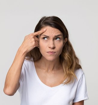 Eine braunhaarige Frau befühlt mit ihrem rechten Zeigefinger ihre Stirn, auf der viele Falten zu sehen sind. Dabei guckt sie skeptisch nach oben, in Richtung ihres Zeigefingers.