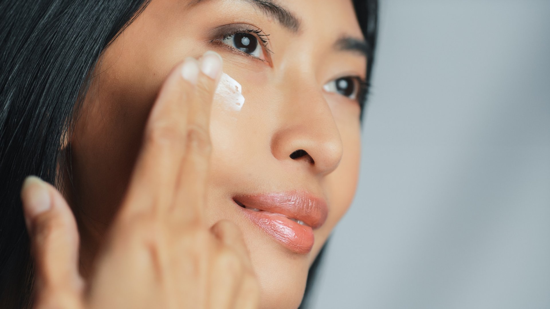 Asiatische Frau streicht sanft mit zwei Fingern Augencreme unter ihr rechtes Auge