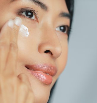 Asiatische Frau streicht sanft mit zwei Fingern Augencreme unter ihr rechtes Auge