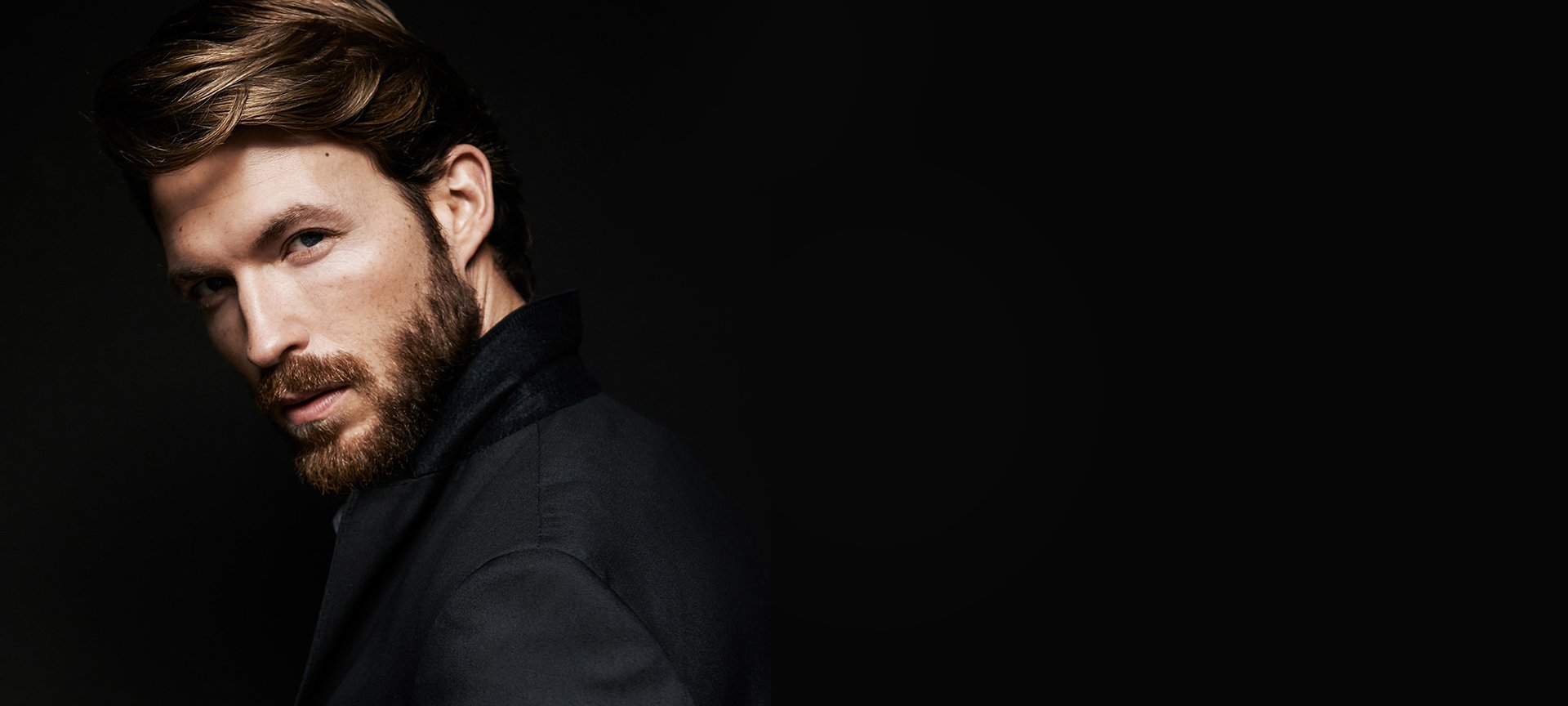 Bärtiger Mann mit Koteletten und schwarzem Jacket vor schwarzem Hintergrund