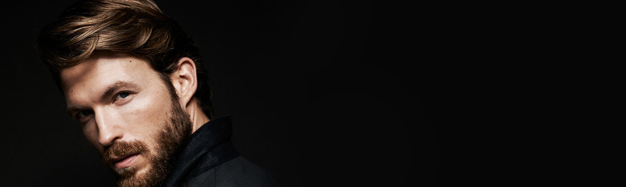 Bärtiger Mann mit Koteletten und schwarzem Jacket vor schwarzem Hintergrund