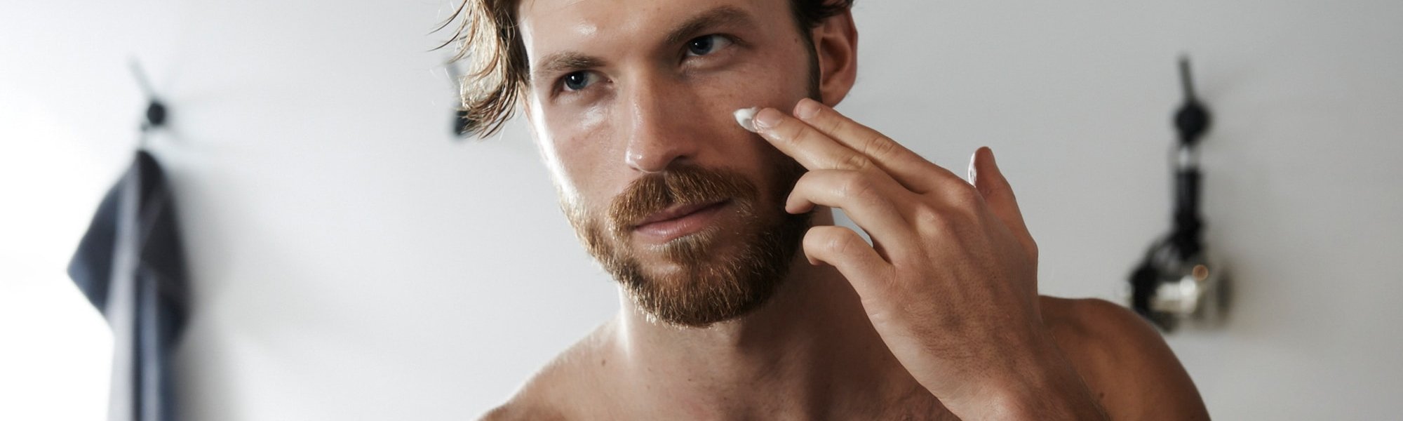 Ein junger Mann betrachtet sich im Spiegel und trägt dabei auf seine Wange eine Creme auf