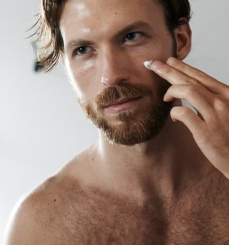 Ein junger Mann betrachtet sich im Spiegel und trägt dabei auf seine Wange eine Creme auf