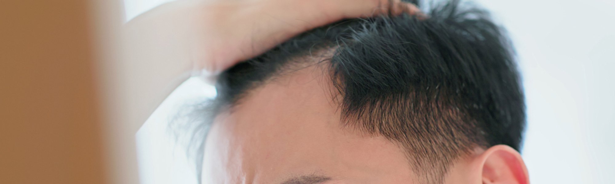 Junger Mann mit schwarzen Haaren und braunen Augen sieht sich seine Geheimratsecken im Spiegel an