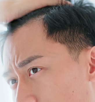 Junger Mann mit schwarzen Haaren und braunen Augen sieht sich seine Geheimratsecken im Spiegel an