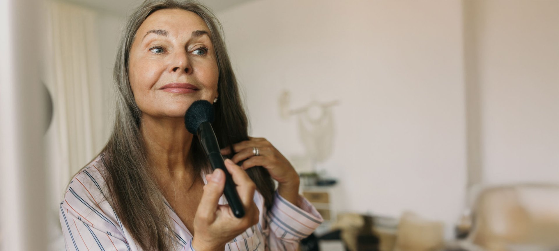 Make-up für reife Haut: Ältere Frau fixiert ihr Make-up mit einem Puderpinsel und betrachtet sich dabei im Spiege