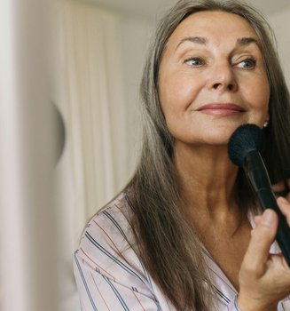 Make-up für reife Haut: Ältere Frau fixiert ihr Make-up mit einem Puderpinsel und betrachtet sich dabei im Spiege