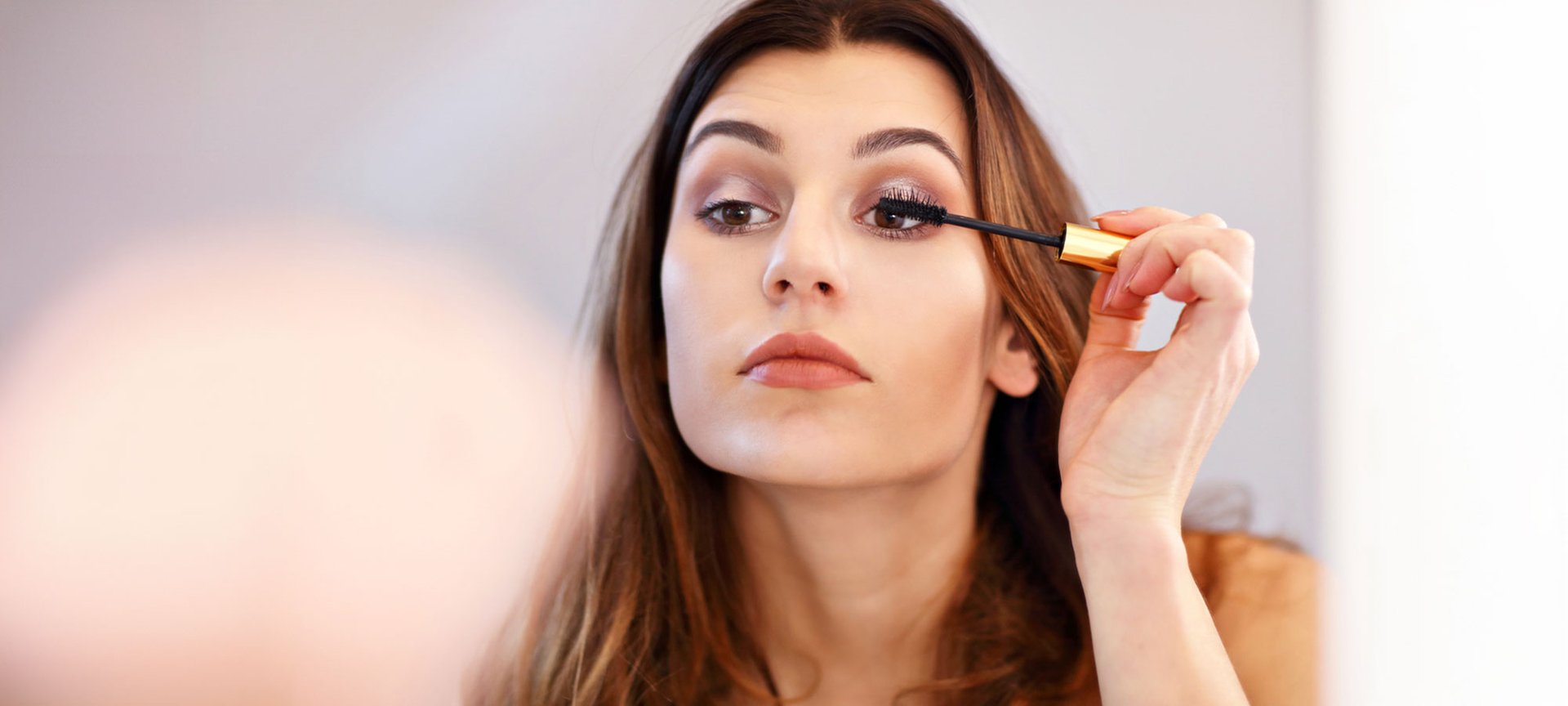 Eine junge Frau mit braunen Haare hält Mascara in der Hand und tuscht die Wimpern ihres linken Auges.