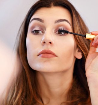 Eine junge Frau mit braunen Haare hält Mascara in der Hand und tuscht die Wimpern ihres linken Auges.
